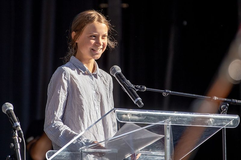 Photo of Greta Thunberg standing behind a podium and microphone smiling with a confident expression