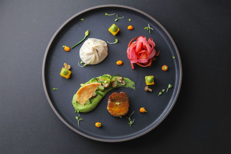 A modern plating on a black plate of crispy pork belly medallion with spring vegetables, avocado, shumai, and pickled rhubarb