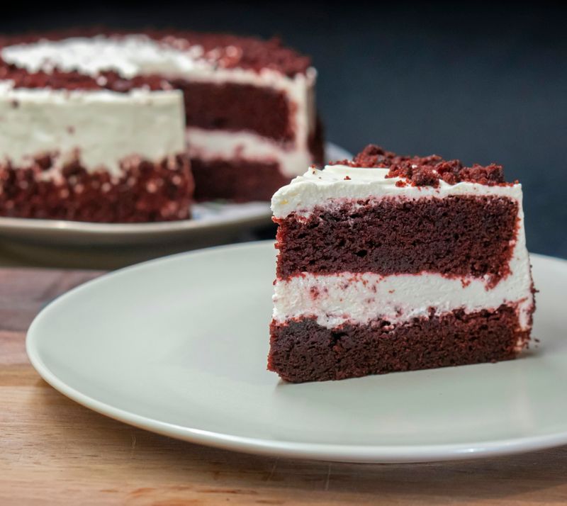 One piece of red velvet cake on a white plate in the foreground with the rest of the whole cake in the background.