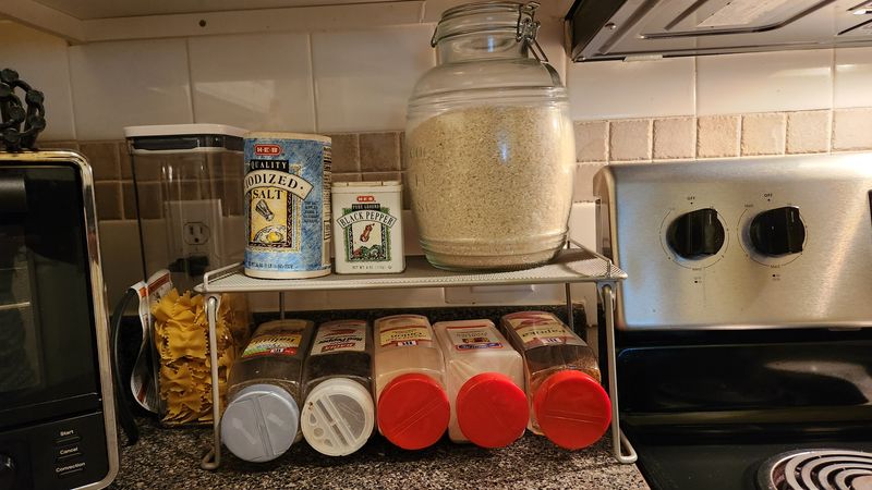 A non-rust shelf beside a stove. It holds spice bottles on the bottom level and other products on top.