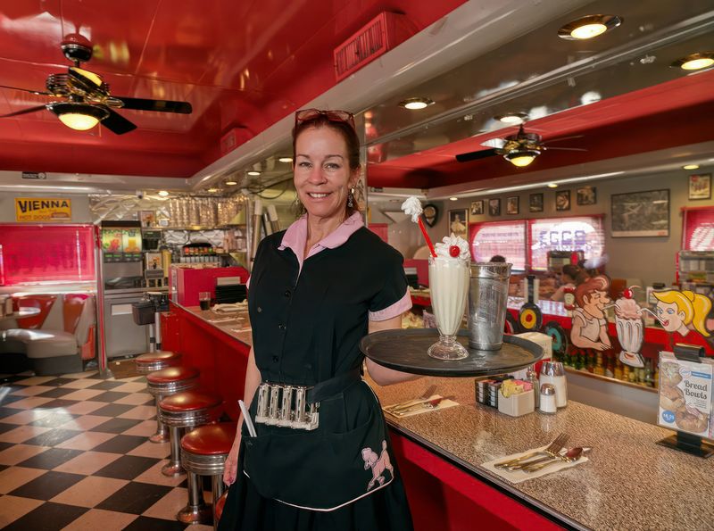 A smiling woman wearing a black server's uniform in a 1950s diner, carrying a tray with a milkshake on it. 