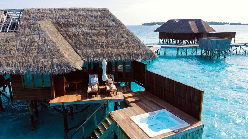 A digital nomad couple relaxing on the deck of an overwater villa above clear blue water. They are working on their laptops.