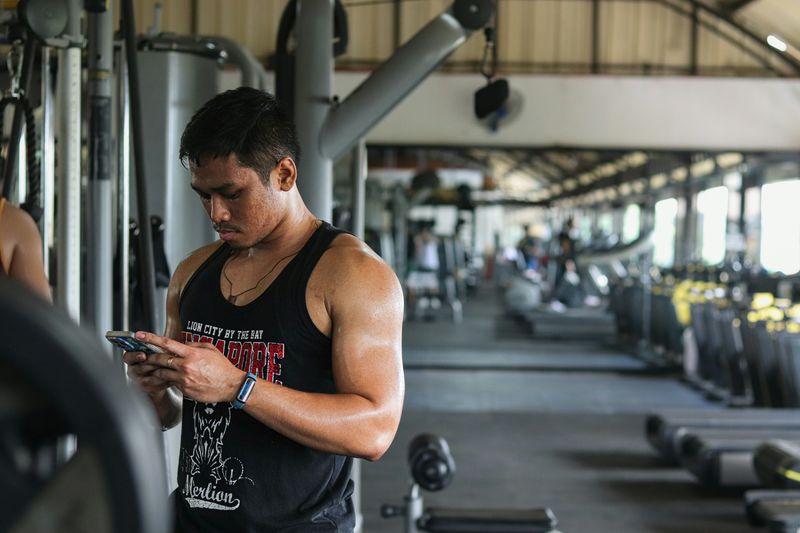 A man stands in a gym looking down at his phone after using a weight machine.
