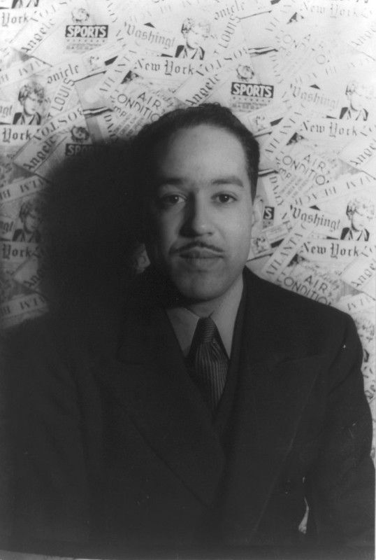 Langston Hughes posing for a photo in front of a wall covered in newspaper clippings.