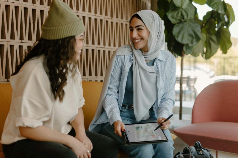 Two women sit in an office and discuss business. One of them holds a tablet with data on it. 