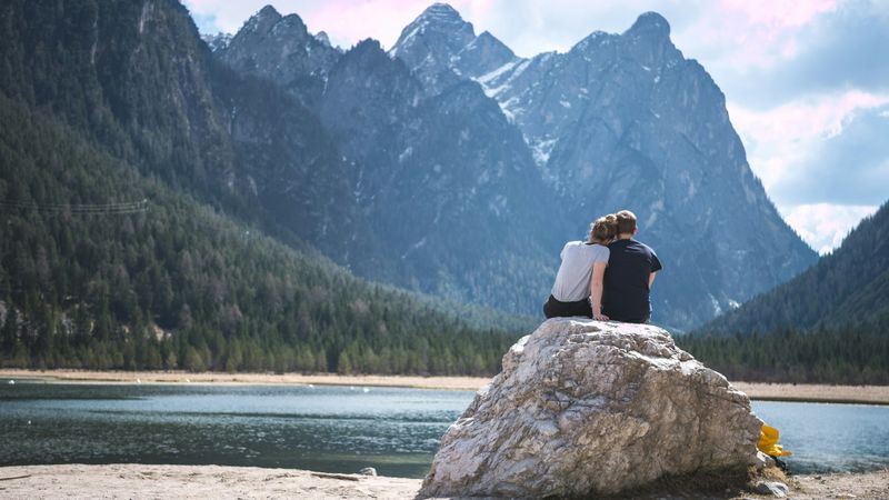 A couple sitting on a rock overlooking a lake and mountains.