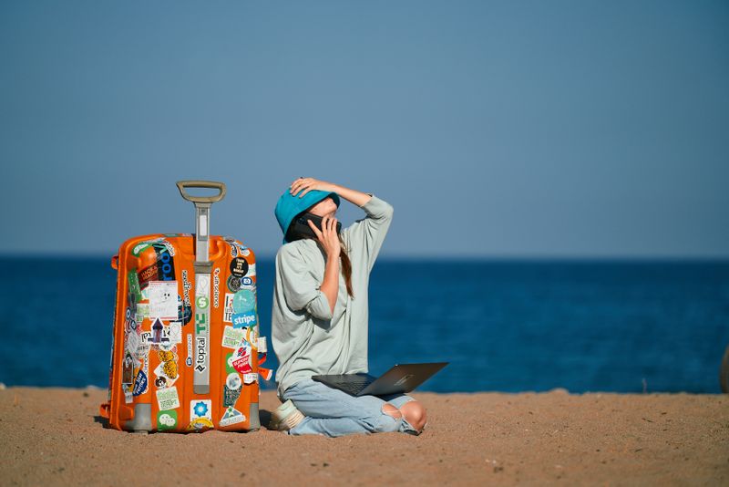 A person on a beach with a laptop and a well used suitcase, talking on their phone while covering their face with their hat.