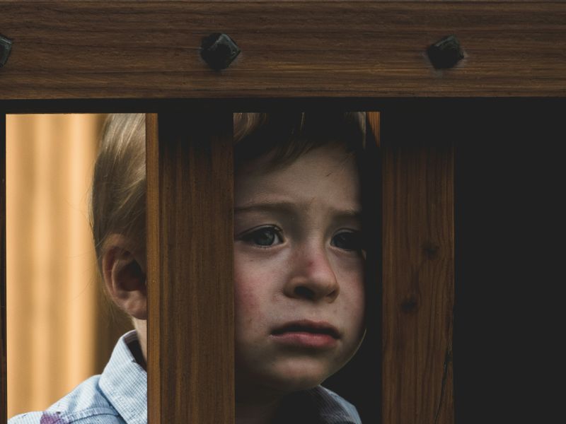 A child with sad face looking through the bars of a wooden fence.