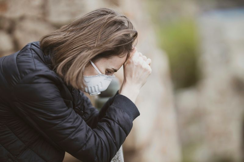 A person feeling sick wearing a medical mask and leaning over with their hands against their head.