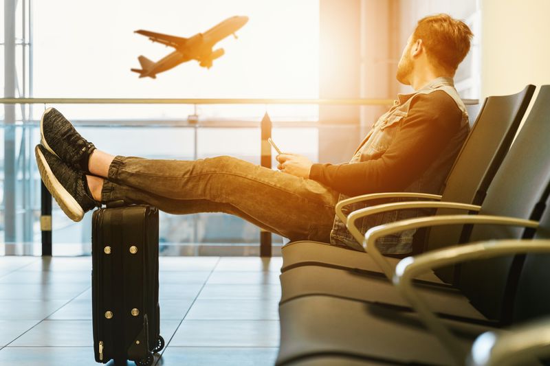 A young man sits in an airport waiting area with his legs up on his carryon, gazing out at an airplane taking off.