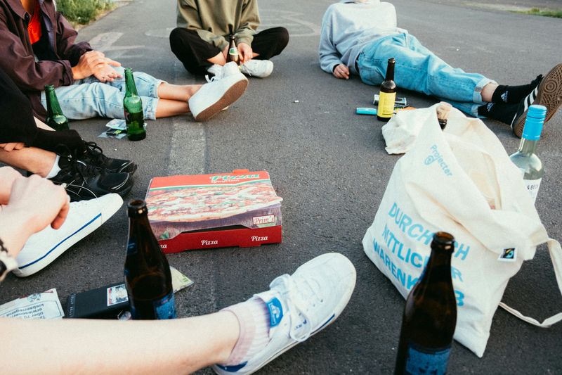 A group sitting on the ground with drinks, a pizza box, and a tote bag nearby.