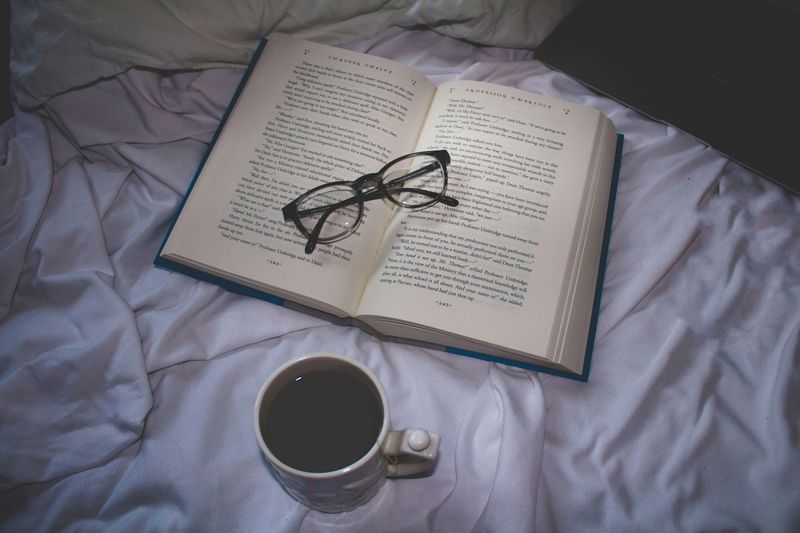 An open book lying on a bed. On top of the pages is a pair of glasses, and near the book is a cup of coffee. 