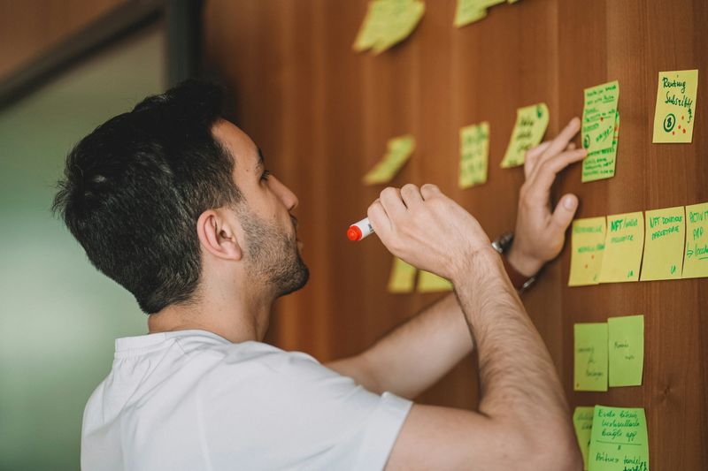 Person with a marker in hand observing many sticky notes with hand-written text attached to a wall. 