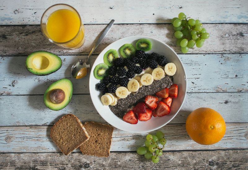 A bowl of fruit, orange juice, avocado, whole wheat bread, grape clusters, and an orange on a wooden table.