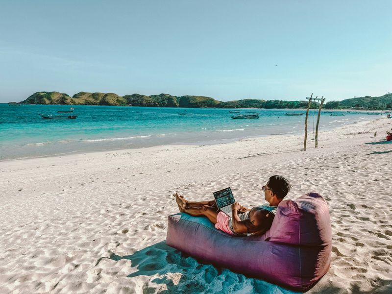 A man sitting on a beanbag sofa on the beach working on his laptop.