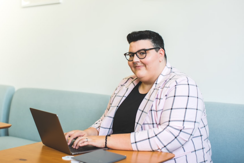 A gender-nonconforming person wearing a blazer, sitting at the computer, with a warm, confident expression.