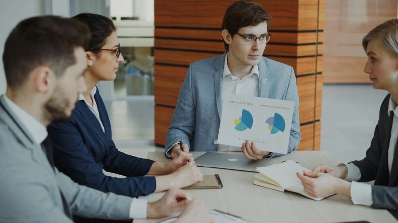 An employee shows a sheet of pie charts to his manager.