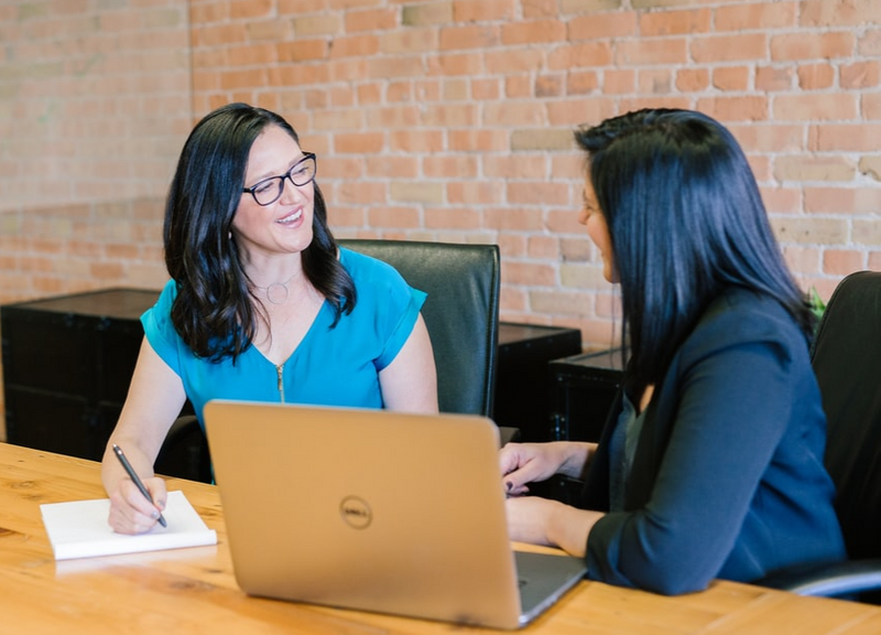 Woman wearing a blazer in discussion with another smiling woman taking notes