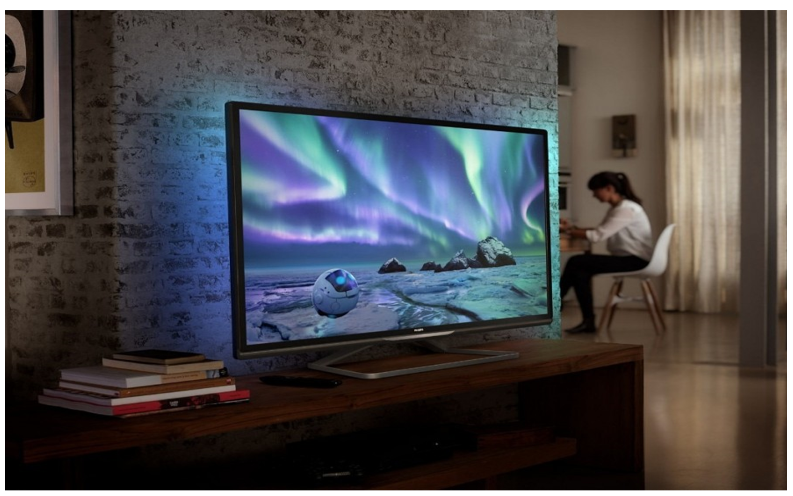 TV on wooden table with books. Screen image of aurora borealis lights and female sits on white chair in background