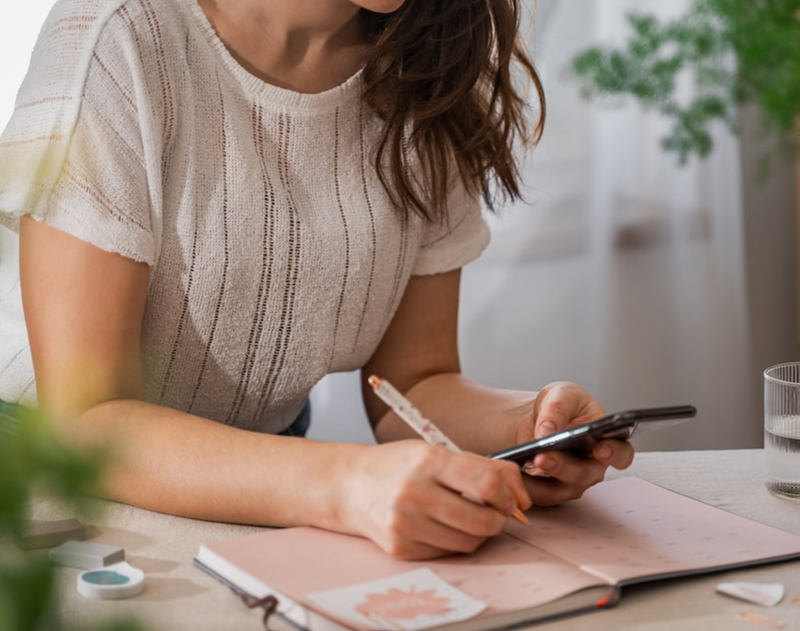 Woman sitting at a desk writing in a book and looking at a phone