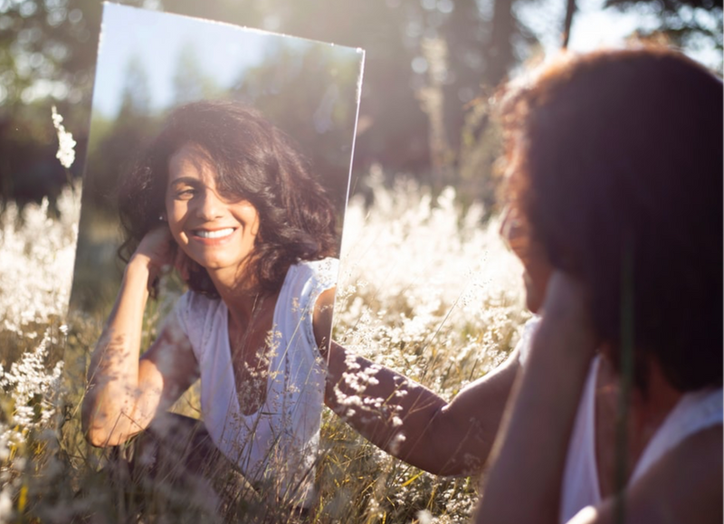 A smiling woman in a field admiring herself in a mirror. 
