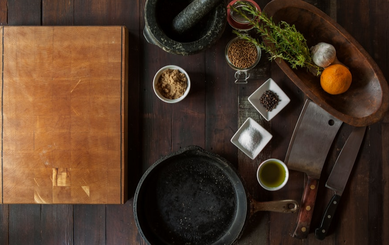 A cutting board, frying pan, mortar & pestle, knives, spices, salt, and fruits on a table.