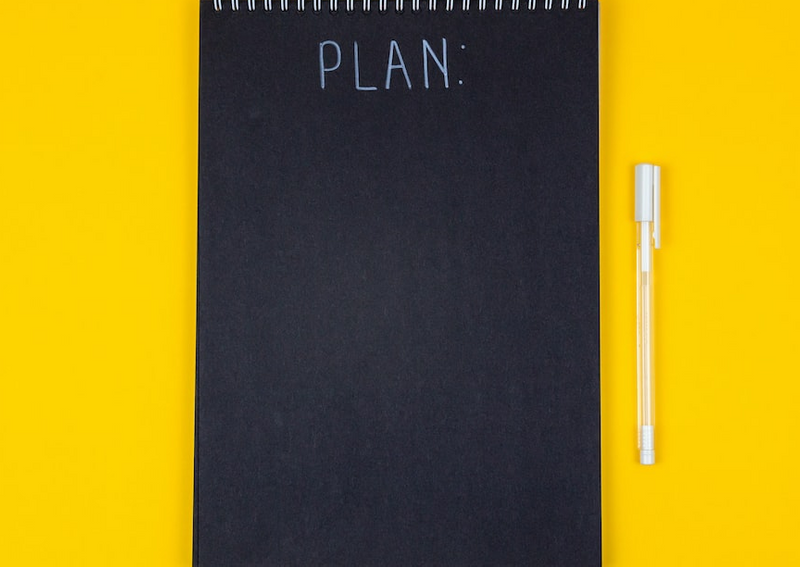 Black notebook with the work 'PLAN' on it on a yellow background