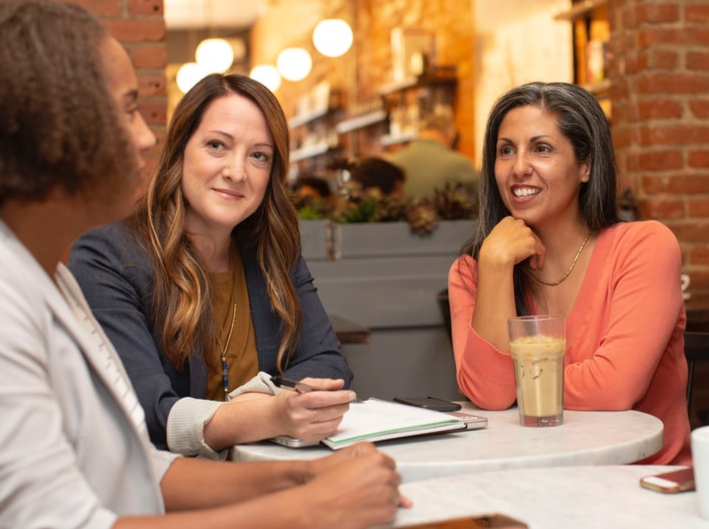 Middle-aged women sitting around a restaurant table, talking, drinking ice coffee, and taking notes.