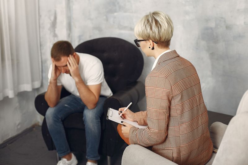 A man sitting in a psychiatrists office showing distress while a female psychiatrist listens and takes notes.