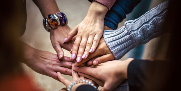 A group of people place their hands together in a gesture of unity and teamwork, with diverse bracelets and sleeves visible.