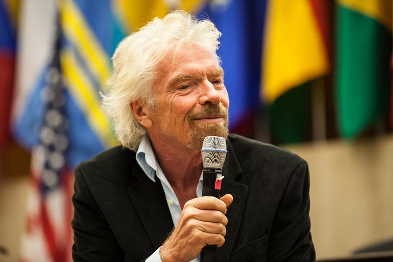 Headshot of Richard Branson in front of a variety of country flags and holding a microphone near his mouth.