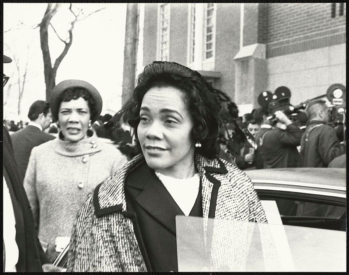 A black and white image of Coretta Scott King beside a car while the press films behind her.