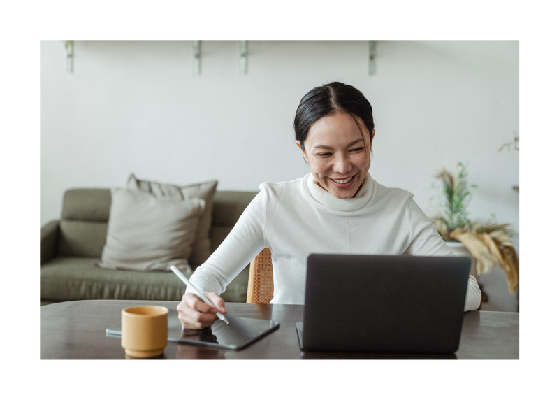 Smiling woman at a home desk writing on notepad looking at a laptop