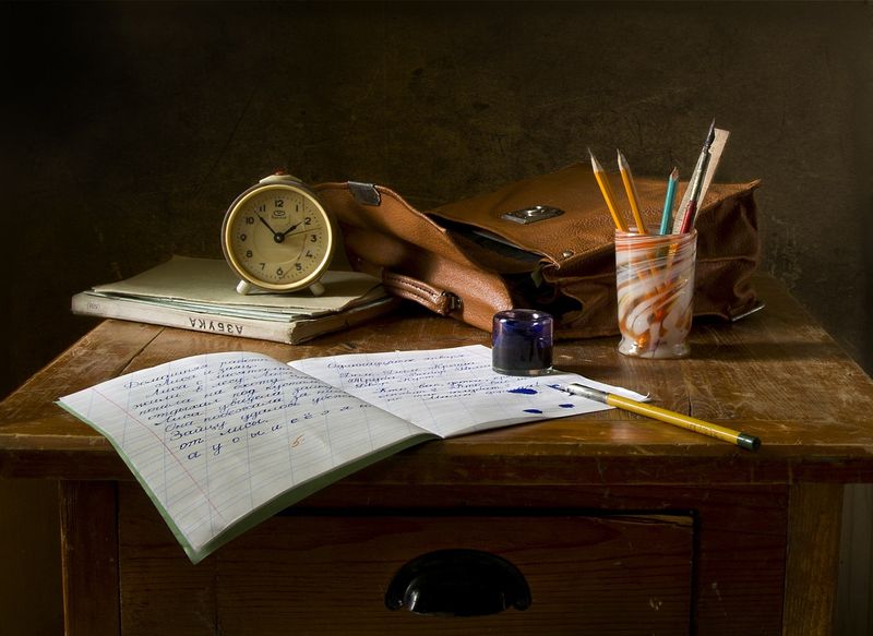 A desk with objects: an open notebook with writing, pen, inkwell, bookbag, papers, clock, and writing utensils.