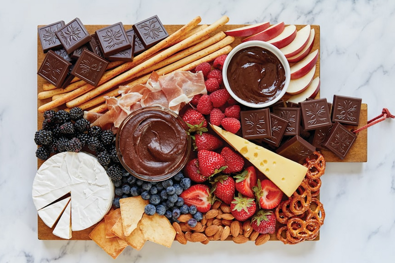 Dessert charcuterie: chocolate, brie cheese, berries, pretzels, long cracker, apples, chocolate dip,  and prosciutto.