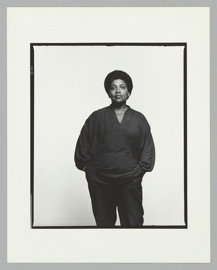 A black & white photo of Audre Lorde posing in front of a white wall.