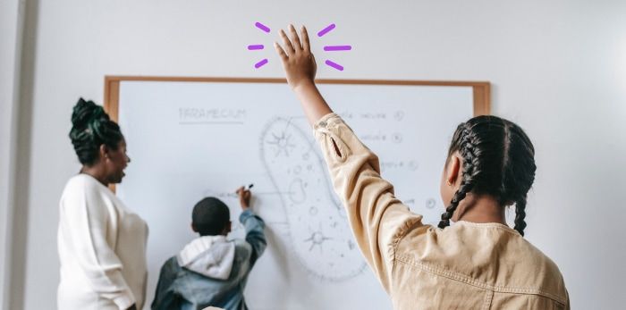 Young boy is at the board answering a question and a young girl is raising her hand as the teacher looks on