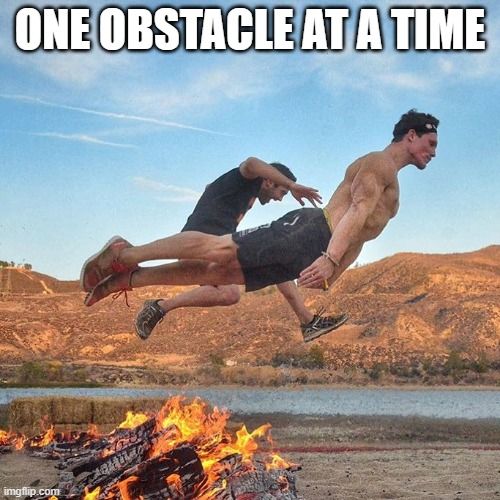 Two men leap over a fire along a lakeshore. The text reads, 'One obstacle at a time.'