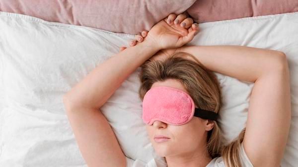 Woman  wearing a pink sleep mask sleeping on her back, with her hands crossed above her head. 