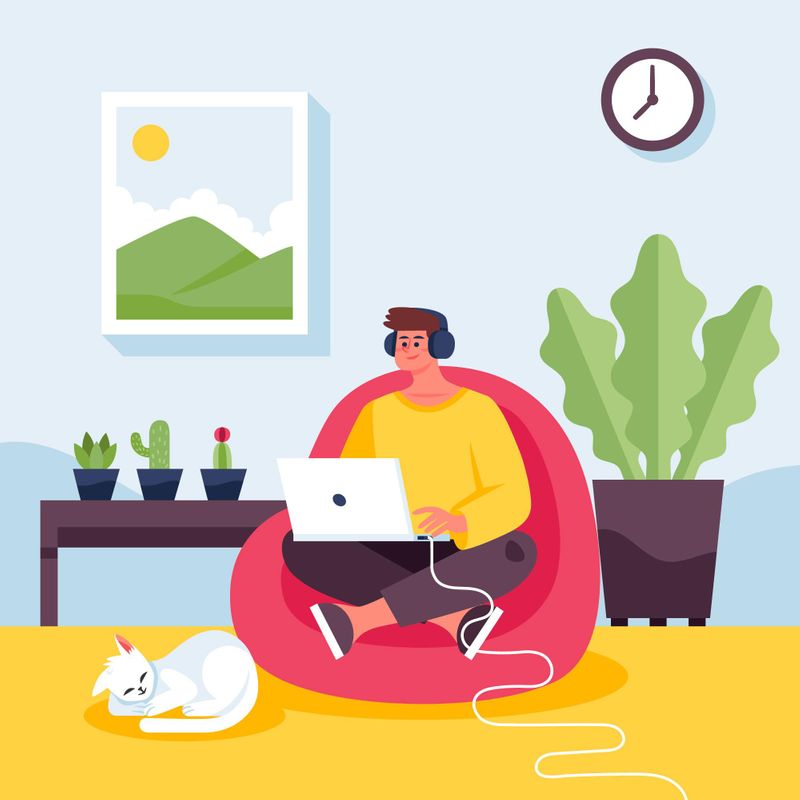 A person sitting on a sofa while working on their laptop in a living room. Their cat sleeps beside them.