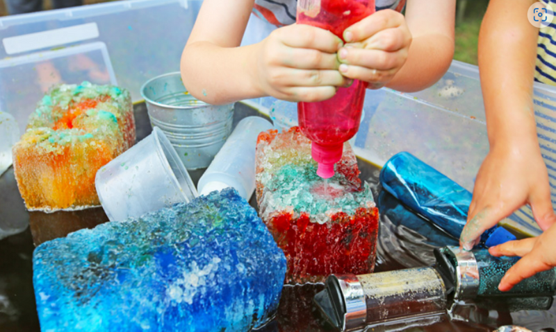 Childrens' hands inside a large tub filled with colored ice blocks and a variety of tools: cups, small buckets, and shakers. 