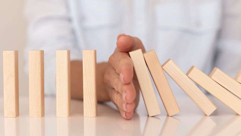A hand stops wooden blocks from toppling like dominoes, symbolizing risk prevention.