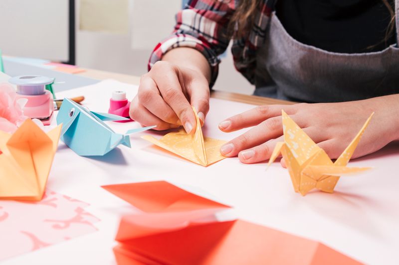 A person's hand carefully folding colorful origami paper cranes on a flat surface.