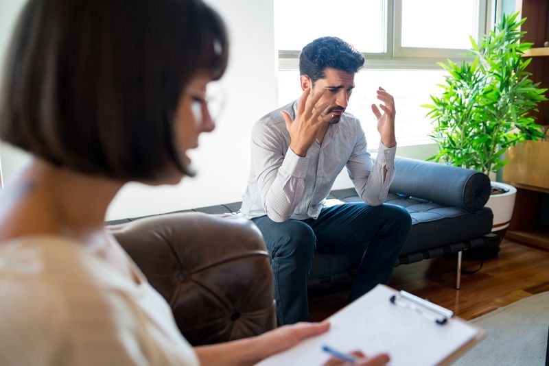 Psychologist taking notes on clipboard in therapy with male patient sitting on sofa