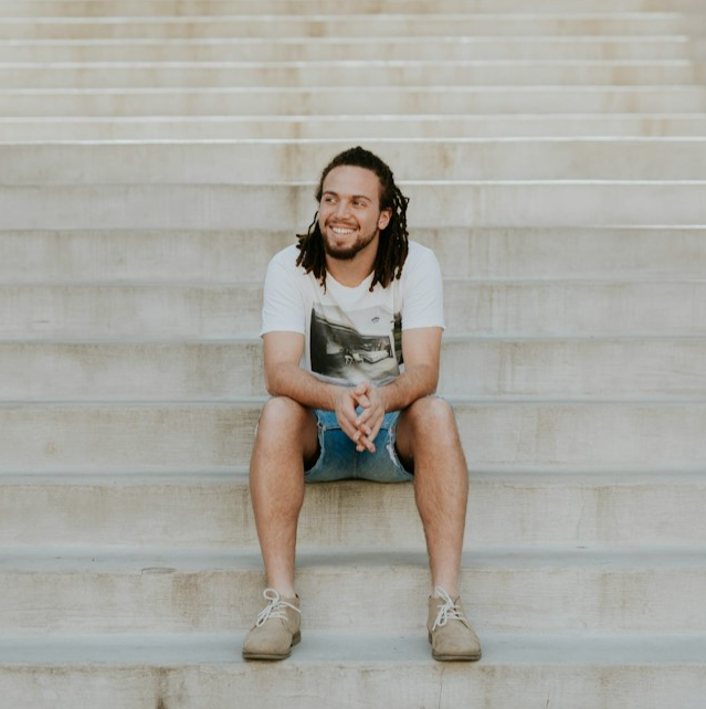 Young man in shorts and short sleeves sitting on steps smiling and looking hopeful