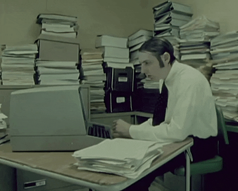 Man surrounded by stacks of paperwork and old, box computer