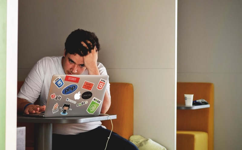 A man is sitting at a desk looking at his laptop screen. His palm is on his head and he looks frustrated.