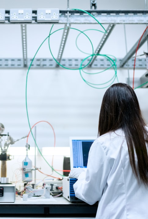 A woman in a lab coat works on the computer at a desk with wires and mechanical equipment.