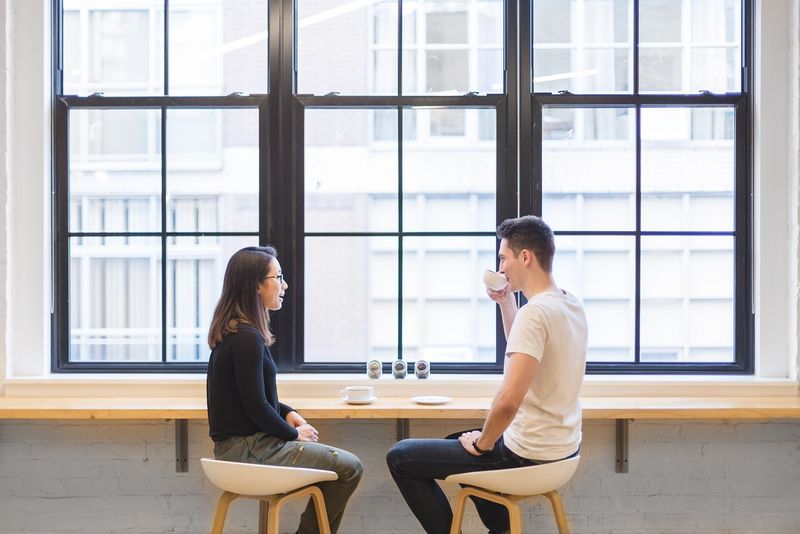 A man and woman sit at a coffee shop drinking drinks in mugs.