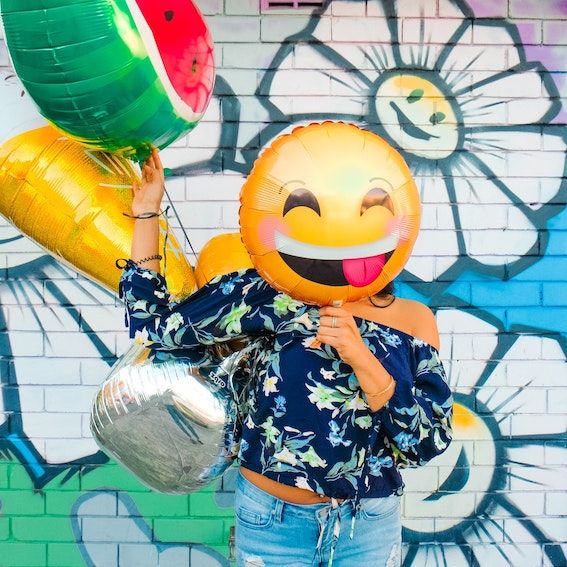 Woman behind a graffiti wall. Holds a smiley faced ballon over of her face. Holds 3 balloons in her left hand.